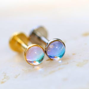 Może przedstawiać: Zbliżenie dwóch kolczyków sztyftów z okrągłymi, iryzującymi kamieniami. Jeden kolczyk ma złoty sztyft, a drugi srebrny. Kamienie prezentują mieszankę odcieni niebieskiego, fioletowego i zielonego.