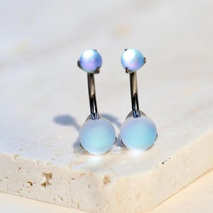 Puede incluir: Dos piercings para el ombligo con orbes iridiscentes de color azul claro y pequeñas gemas de tono púrpura. La joyería tiene postes plateados. Los anillos se exhiben sobre una superficie clara y texturizada.