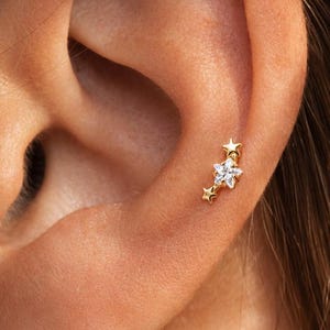 May include: A gold-toned ear stud featuring a cluster of three stars arranged in a vertical line. The central star is larger and set with a clear, sparkling gemstone, while the two smaller stars at the top and bottom are plain gold metal.