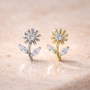 May include: Two flower-shaped stud earrings. One earring is silver-toned, the other is gold-toned. Each earring features a central, round, clear gemstone surrounded by petals. The stems have two leaf-shaped accents.