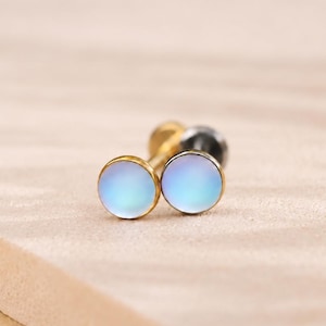 May include: A pair of stud earrings featuring circular, iridescent, light blue cabochon stones set in gold-toned metal. The earrings have a flat, smooth surface and a metallic post backing, showcasing a reflective, opalescent finish on the front.