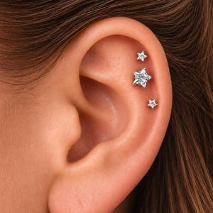 May include: Three silver-toned star-shaped stud earrings are arranged vertically along the upper cartilage of the ear. Each star features a central clear gemstone, with the middle star being the largest of the three pieces.