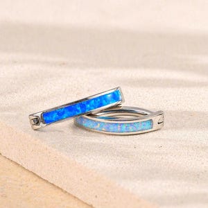 May include: Two silver hoop earrings with a vibrant blue opal inlay. The earrings are displayed on a light beige surface, showcasing their bright color and design. The opal has a shimmering, iridescent quality.
