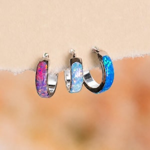 May include: Three small hoop earrings featuring inlaid opal stones. The left hoop has a pink and purple opal inlay, the center hoop has a light blue opal inlay, and the right hoop has a vibrant blue opal inlay, all set in a polished silver-tone metal.