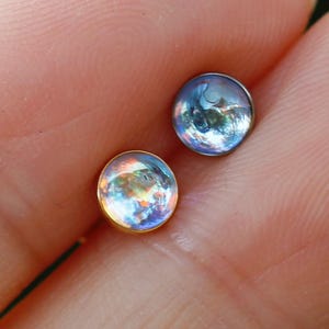 May include: Two small circular stud earrings with iridescent, dome-shaped cabochons. One earring features a gold-toned metal setting, while the other is set in a dark, cool-toned metal. Both stones display a reflective, multi-colored sheen.