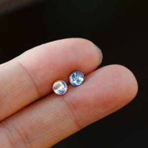 May include: Two small, round stud earrings featuring iridescent, translucent cabochon stones. One earring is set in a warm gold-toned metal, while the other is set in a cool silver-toned metal, both displaying subtle blue and white light reflections.