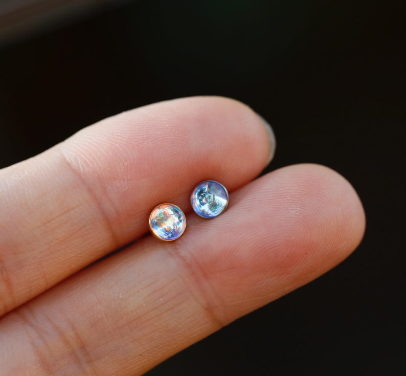 May include: Two small, round stud earrings featuring iridescent, translucent cabochon stones. One earring is set in a warm gold-toned metal, while the other is set in a cool silver-toned metal, both displaying subtle blue and white light reflections.