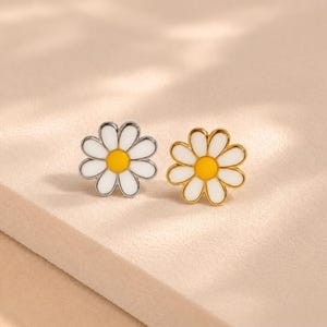 May include: Two daisy-shaped stud earrings, one with a silver-toned metal border and the other with a gold-toned metal border. Both earrings feature white enamel petals surrounding a bright yellow enamel center, creating a floral design.