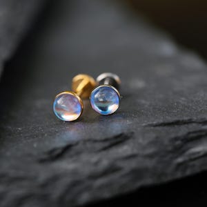 May include: Two stud earrings featuring round, translucent, iridescent cabochon stones that display a soft blue and pink glow. The earrings have metallic posts and are positioned side by side, showcasing their smooth, domed surfaces.