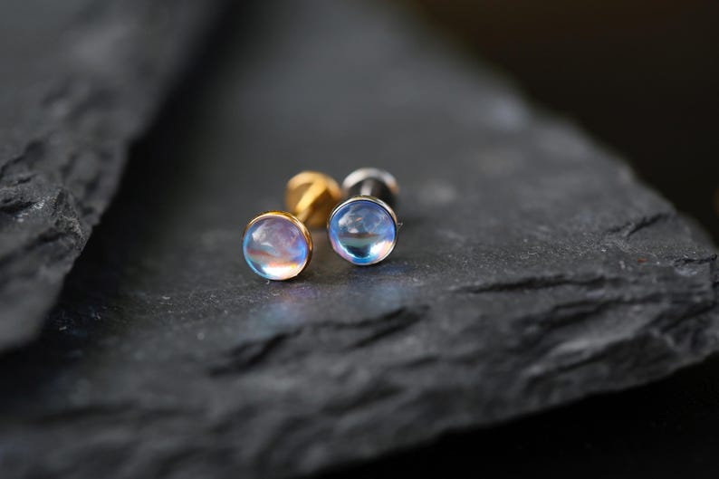May include: Two stud earrings featuring round, translucent, iridescent cabochon stones that display a soft blue and pink glow. The earrings have metallic posts and are positioned side by side, showcasing their smooth, domed surfaces.