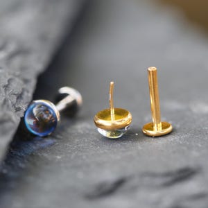 May include: Three stud earrings with flat back posts. One earring features a round, iridescent blue stone. Two other earrings have gold-toned metal posts and settings, with one showing the back post and the other showing a clear, rounded stone top.