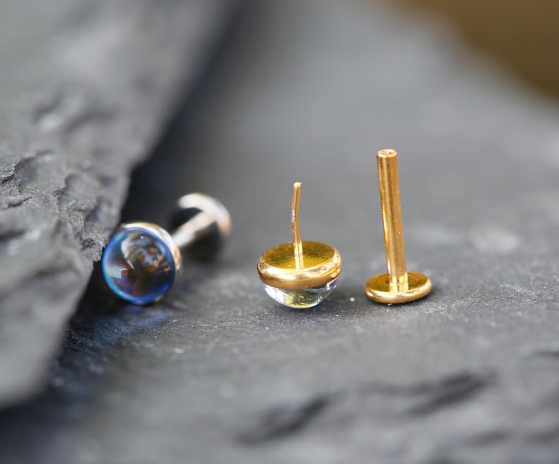 May include: Three stud earrings with flat back posts. One earring features a round, iridescent blue stone. Two other earrings have gold-toned metal posts and settings, with one showing the back post and the other showing a clear, rounded stone top.