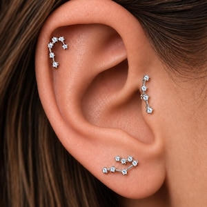 May include: Three silver-toned ear piercings featuring small, clear, round-cut stones arranged in linear, constellation-like patterns. One piece is located on the upper cartilage, one on the tragus, and one on the lower earlobe.
