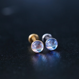 May include: Two flat-back stud earrings featuring round, iridescent cabochon stones that display a soft blue and pink glow. One earring has a gold-toned metal base, while the other features a silver-toned metal base, both with a smooth finish.