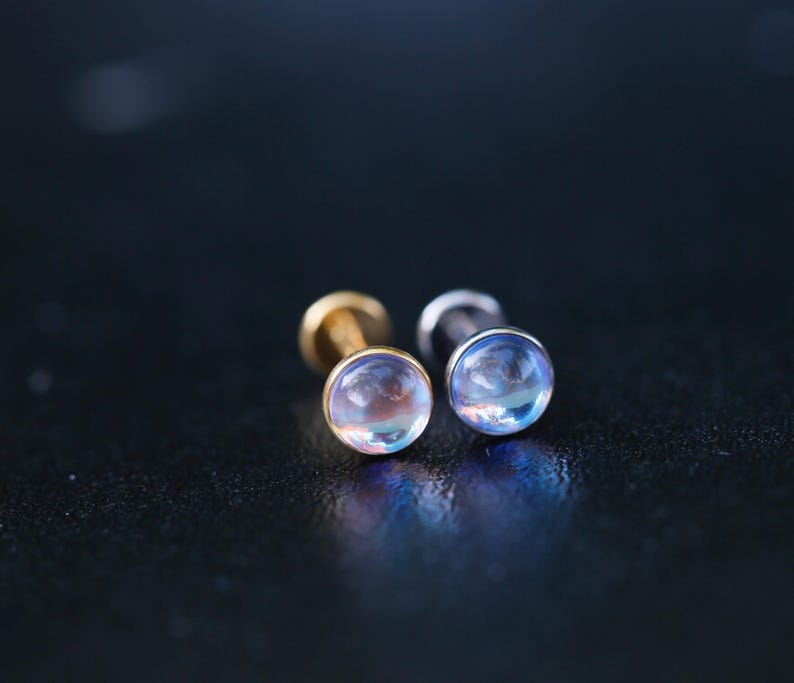 May include: Two flat-back stud earrings featuring round, iridescent cabochon stones that display a soft blue and pink glow. One earring has a gold-toned metal base, while the other features a silver-toned metal base, both with a smooth finish.