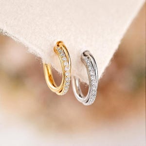 May include: Two small hoop earrings, one in gold and one in silver, each featuring a double-band design where one band is smooth and the other is encrusted with a row of small, clear, sparkling stones set along the front surface.