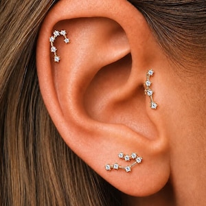 May include: Three gold-toned ear piercings featuring small, clear, round-cut stones arranged in constellation-like patterns. One piece is positioned on the upper cartilage, one on the mid-ear cartilage, and one on the lower earlobe.