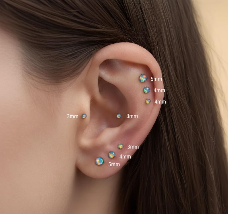 May include: A collection of round opal stud earrings in various sizes, including 3mm, 4mm, and 5mm, displayed in multiple ear piercings. Each earring features a iridescent, multi-colored opal stone set in a simple metal base, showing clear size labels.