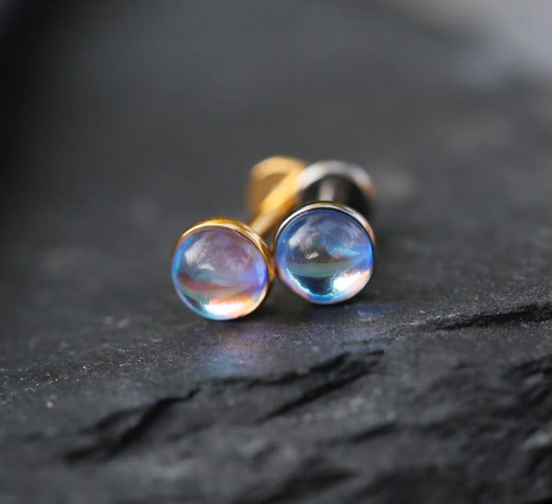 May include: A pair of stud earrings featuring round, iridescent cabochon stones with a blue and pink glow, set in gold-toned metal posts. The stones have a smooth, domed surface that reflects light, creating a soft, ethereal appearance on the jewelry.