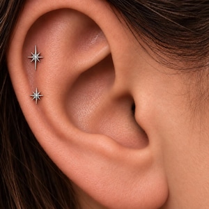 May include: Two silver-toned starburst-shaped earrings are positioned vertically along the upper cartilage of the ear. Each earring features a central point with radiating spikes, creating a symmetrical, celestial-inspired design on the ear.
