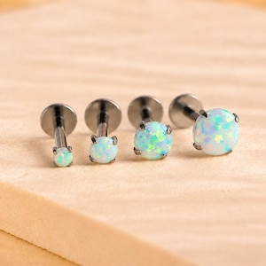 May include: Four labret stud earrings with dark metal posts and flat back discs. Each earring features a round, prong-set white opal stone with iridescent blue and green flashes. The stones vary in size, arranged in a row from smallest to largest.
