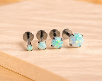 Puede incluir: Cuatro piercings labret con postes de metal oscuro y bases circulares planas. Cada pieza presenta una piedra de ópalo blanco redonda engastada con garras, con destellos iridiscentes azules y verdes, aumentando de tamaño de izquierda a derecha.
