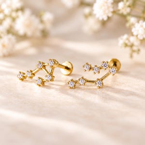 May include: A pair of gold-toned stud earrings featuring a constellation design. Each earring is crafted with small, clear, round-cut stones set in a metal frame that mimics a star pattern, complete with a flat circular backing post for secure wear.