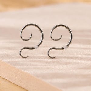 May include: A pair of silver-toned metal ear cuffs featuring a continuous spiral design. Each piece has a smooth, curved wire structure that forms a large upper loop and a smaller lower hook, with a small white silicone sleeve on the lower section.