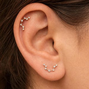 May include: Two gold-toned earrings featuring a constellation design with small, clear, round-cut stones set along thin metal bars. One earring is positioned on the upper cartilage, while the other is placed on the earlobe.