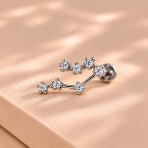 以下が含まれることがあります： 星座をモチーフにしたシルバー調のピアス。曲線を描く金属フレームに、7つの小さな透明なラウンドカットストーンがセットされています。裏側には平らな円形の留め具が付いた、繊細で構造的なジュエリーアイテムです。