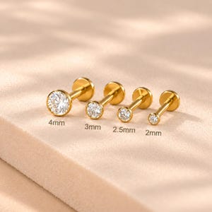 Könnte beinhalten: Vier goldfarbene Labret-Stecker mit klaren, rund geschliffenen Steinen in Zargenfassung. Die Stecker sind nach Größe sortiert, beschriftet mit 4mm, 3mm, 2,5mm und 2mm, und verfügen über flache Rückplatten sowie gerade Stäbe für Piercings.
