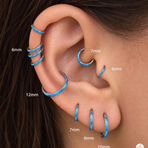 May include: A collection of silver-toned hoop earrings with blue inlay, featuring various sizes labeled as 6mm, 7mm, 8mm, 10mm, and 12mm. These circular earrings are displayed in multiple ear piercings, showcasing a consistent blue and silver aesthetic.