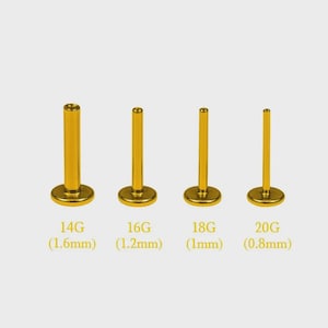Op de afbeelding: Vier goudkleurige labret-piercings met een platte achterkant, gerangschikt in afnemende dikte. De tekst onder elke piercing geeft de dikte en maat aan: 14G (1,6 mm), 16G (1,2 mm), 18G (1 mm) en 20G (0,8 mm). Elke piercing heeft een ronde voet.