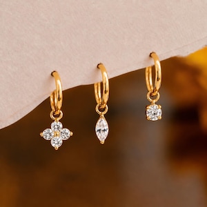 May include: Three gold hoop earrings with dangling charms. The first features a four-petaled flower shape with clear stones, the second has a marquise-cut clear stone, and the third displays a single round clear stone. All are set in gold metal.