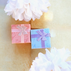 May include: Two square gift boxes, one in a textured coral pink and the other in a textured light blue, each adorned with a matching satin ribbon tied in a bow on top. The boxes are positioned between two large, white, fluffy decorative flowers.