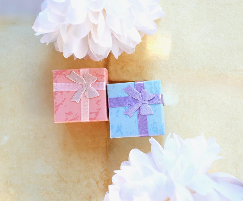 May include: Two square gift boxes, one in a textured coral pink and the other in a textured light blue, each adorned with a matching satin ribbon tied in a bow on top. The boxes are positioned between two large, white, fluffy decorative flowers.