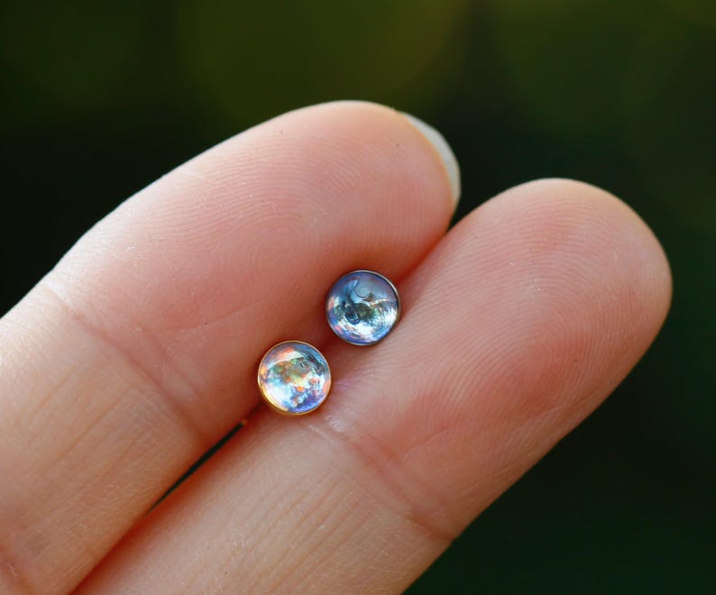 May include: Two small circular stud earrings with iridescent, dome-shaped cabochons. One earring features a gold-toned metal setting, while the other is set in a dark, cool-toned metal. Both stones display a reflective, multi-colored sheen.