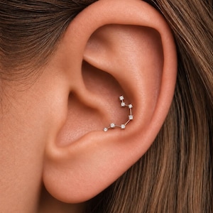 May include: A silver-toned ear piercing jewelry piece featuring a curved design adorned with six small, clear, round-cut stones arranged in a constellation-like pattern, securely positioned within the conch area of the ear cartilage.