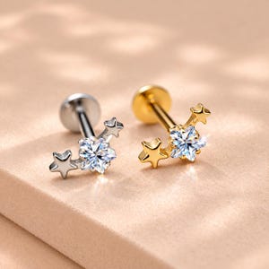 May include: Two flat-back labret studs, one silver-toned and one gold-toned, each featuring a central clear faceted stone shaped like a star, flanked by two smaller solid metal stars on either side of the main setting, creating a celestial design.
