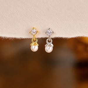 May include: Two stud earrings, one gold-toned and one silver-toned, each featuring a clear round-cut gemstone at the top and a small white pearl drop suspended from a metal cap. The earrings are displayed side by side against a light background.