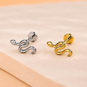 May include: Two flat back stud earrings shaped like snakes, one in a polished silver tone and one in a polished gold tone. Each earring features a detailed snake design with a curved body and a flat circular backing for secure placement.