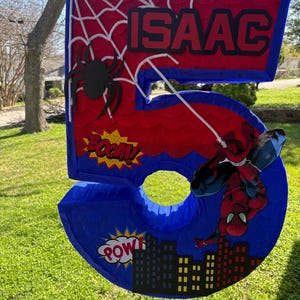 K&ouml;nnte beinhalten: Eine gro&szlig;e, rote und blaue Pi&ntilde;ata mit der Zahl 5 und einem Spider-Man-Thema. Die Pi&ntilde;ata tr&auml;gt den Namen "ISAAC", ein Spinnennetz-Design und Comic-Textblasen mit "BOOM" und "POW".