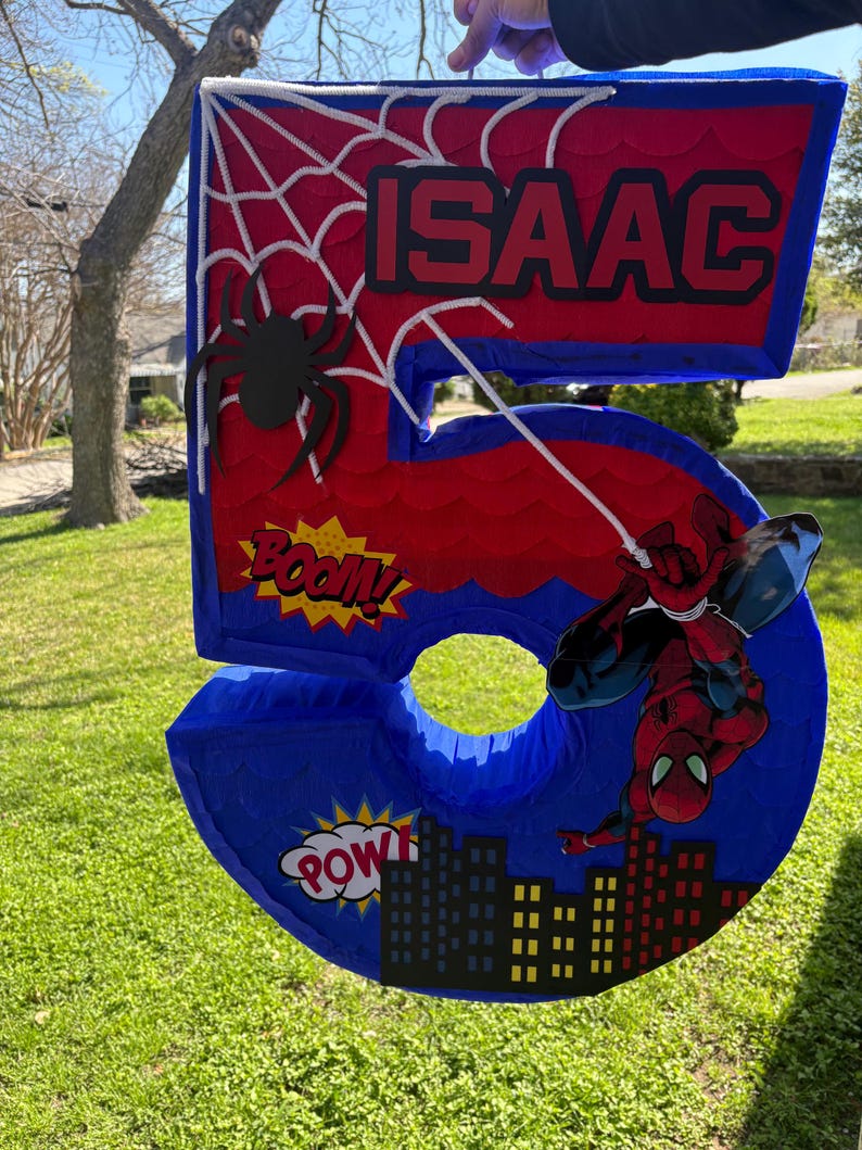 K&ouml;nnte beinhalten: Eine gro&szlig;e, rote und blaue Pi&ntilde;ata mit der Zahl 5 und einem Spider-Man-Thema. Die Pi&ntilde;ata tr&auml;gt den Namen "ISAAC", ein Spinnennetz-Design und Comic-Textblasen mit "BOOM" und "POW".