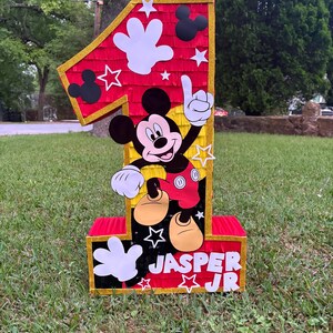 May include: A large, colorful number one pi&ntilde;ata featuring Mickey Mouse. The pi&ntilde;ata is red, black, yellow, and white, with gold trim. The name "JASPER JR" is displayed on the front. The pi&ntilde;ata is decorated with stars, Mickey Mouse hands, and Mickey Mouse silhouettes.