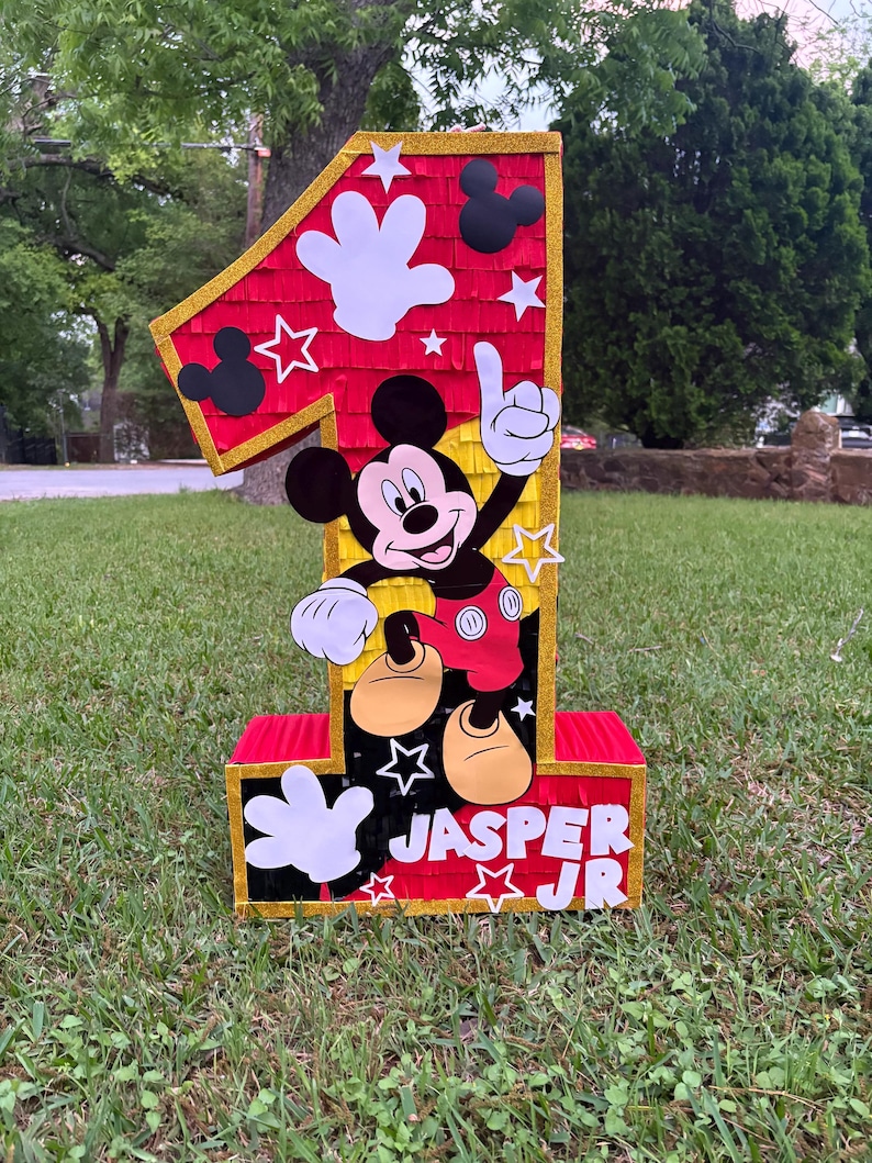 May include: A large, colorful number one pi&ntilde;ata featuring Mickey Mouse. The pi&ntilde;ata is red, black, yellow, and white, with gold trim. The name "JASPER JR" is displayed on the front. The pi&ntilde;ata is decorated with stars, Mickey Mouse hands, and Mickey Mouse silhouettes.