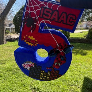 K&ouml;nnte beinhalten: Eine gro&szlig;e, blaue Zahl f&uuml;nf Pi&ntilde;ata mit roten Akzenten, einem Spinnennetz-Design und dem Namen "ISAAC". Mit einer Spider-Man-Grafik und Comic-Text. Ideal f&uuml;r eine Geburtstagsfeier.