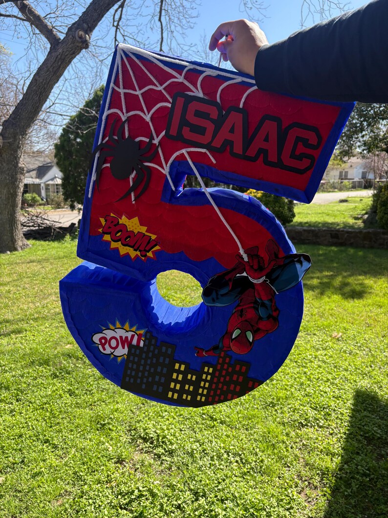 K&ouml;nnte beinhalten: Eine gro&szlig;e, blaue Zahl f&uuml;nf Pi&ntilde;ata mit roten Akzenten, einem Spinnennetz-Design und dem Namen "ISAAC". Mit einer Spider-Man-Grafik und Comic-Text. Ideal f&uuml;r eine Geburtstagsfeier.