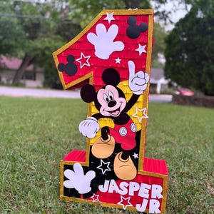 May include: A large, red number one pi&ntilde;ata with gold trim, featuring Mickey Mouse and white hand cutouts. The pi&ntilde;ata has the name "Jasper Jr" on the bottom. Perfect for a first birthday party.