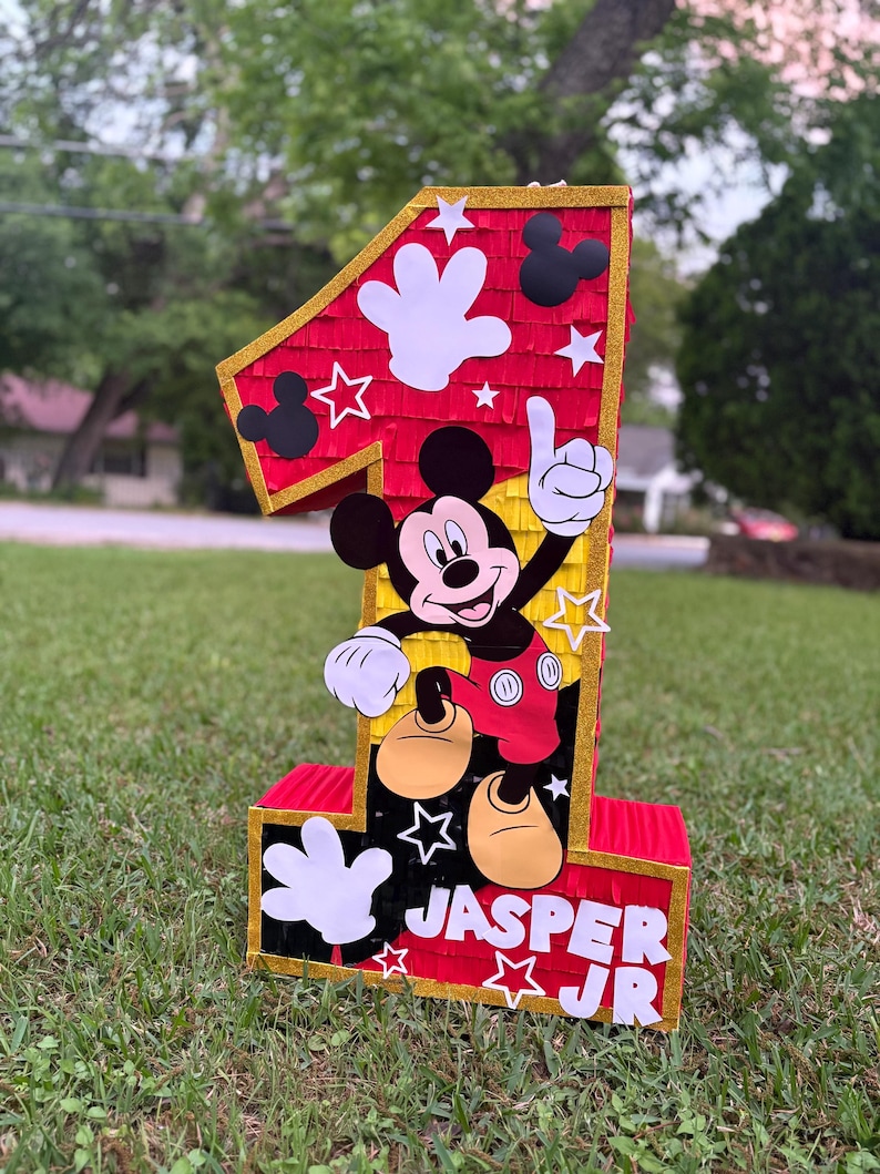 May include: A large, red number one pi&ntilde;ata with gold trim, featuring Mickey Mouse and white hand cutouts. The pi&ntilde;ata has the name "Jasper Jr" on the bottom. Perfect for a first birthday party.