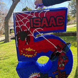 K&ouml;nnte beinhalten: Eine blaue, rote und wei&szlig;e Pi&ntilde;ata mit der Nummer 5 im Spider-Man-Design. Die Pi&ntilde;ata hat ein Spinnennetz-Design, eine schwarze Spinne und den Namen "ISAAC" in Rot. Comic-Soundeffekte und eine Skyline sind ebenfalls enthalten.